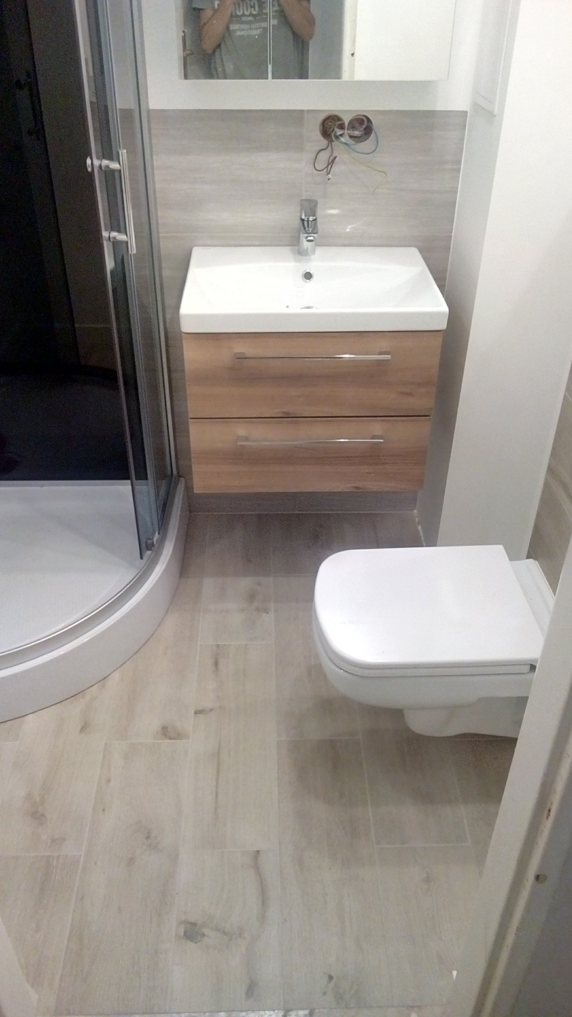 Nowoczesna łazienka z kabiną prysznicową z zaokrąglonym szkłem, umywalką z drewnianą szafką i wiszącą toaletą, wykończona płytkami imitującymi drewno. W tle widoczne przewody elektryczne wystające...