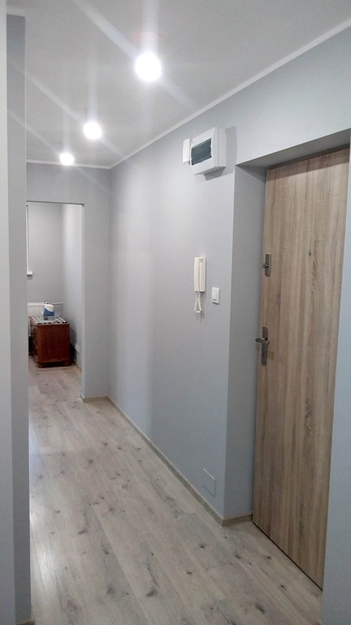 Korytarz po remoncie z szarymi ścianami, panelami podłogowymi imitującymi drewno i drewnianymi drzwiami. Na ścianie widoczny biały domofon i skrzynka elektryczna.