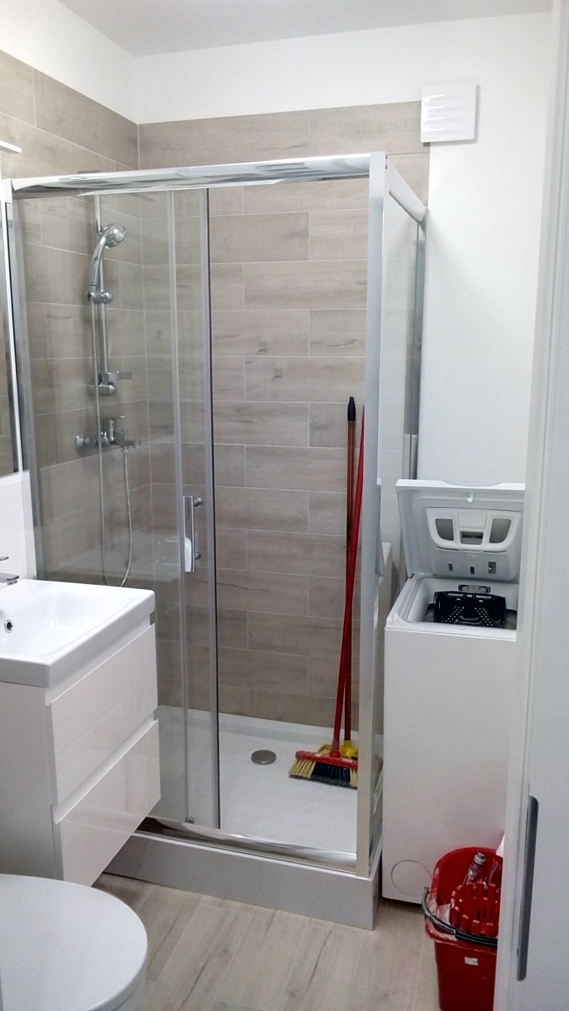 Nowoczesna łazienka z kabiną prysznicową z przesuwnymi drzwiami, białą umywalką z szafką, pralką z otwartym bębnem oraz akcesoriami do sprzątania w rogu kabiny.