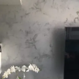 Ściana z dekoracyjną, szarą strukturą przypominającą marmur, nad nią biały klimatyzator, poniżej biała komoda z orchideą w kwadratowej doniczce i czarnym soundbarem.