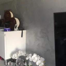 Ściana w odcieniach szarości z efektem dekoracyjnym, obok biała szafka z dekoracjami i kwiatami w białej doniczce, fragment telewizora po prawej stronie.