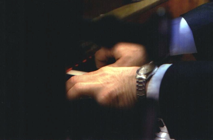 Dłonie mężczyzny w garniturze grającego na pianinie, widoczny zegarek na nadgarstku, ujęcie z bliska w ciemnym otoczeniu.