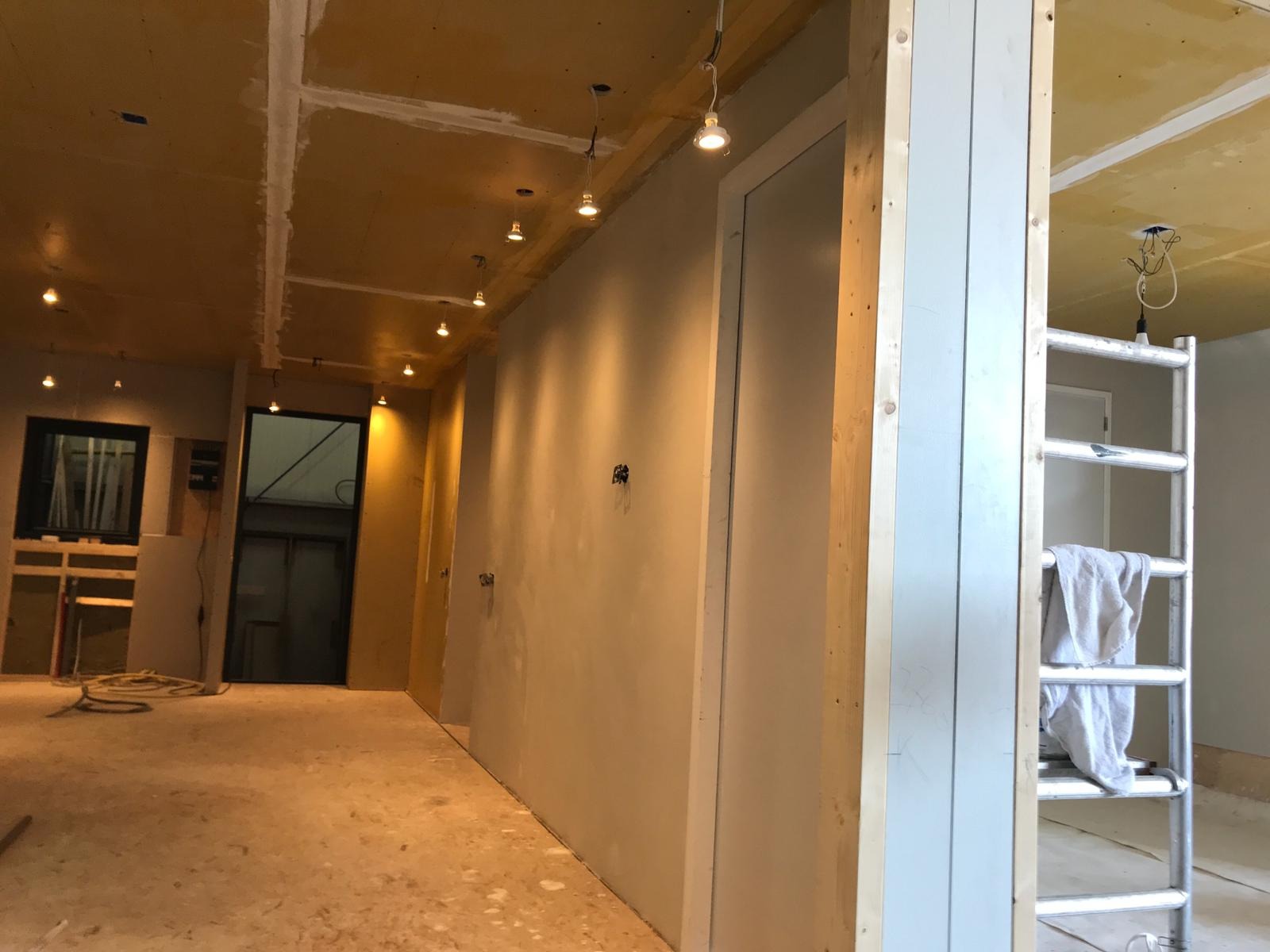Wnętrze w trakcie remontu: surowy sufit z punktowymi lampami, świeżo otynkowane ściany, aluminiowa drabina z ręcznikiem w tle.