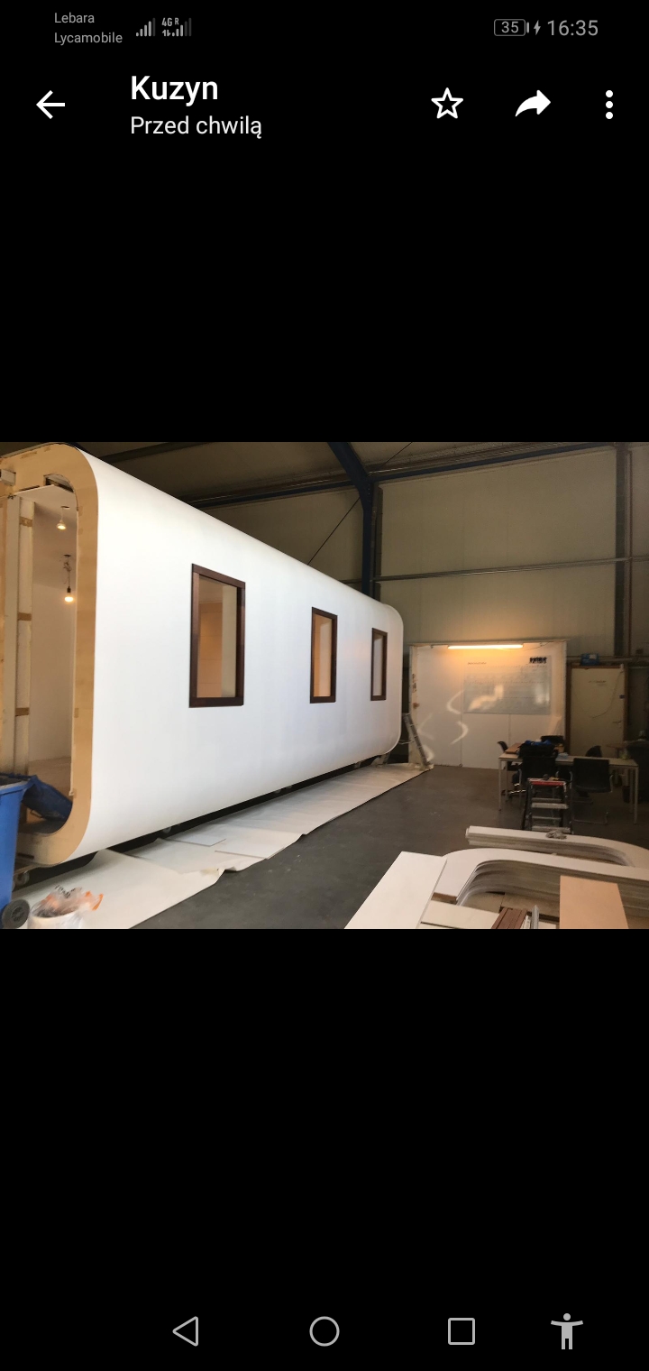 Modułowy, biały dom z brązowymi ramami okiennymi, w trakcie budowy w hali, z widocznymi elementami konstrukcyjnymi i narzędziami.