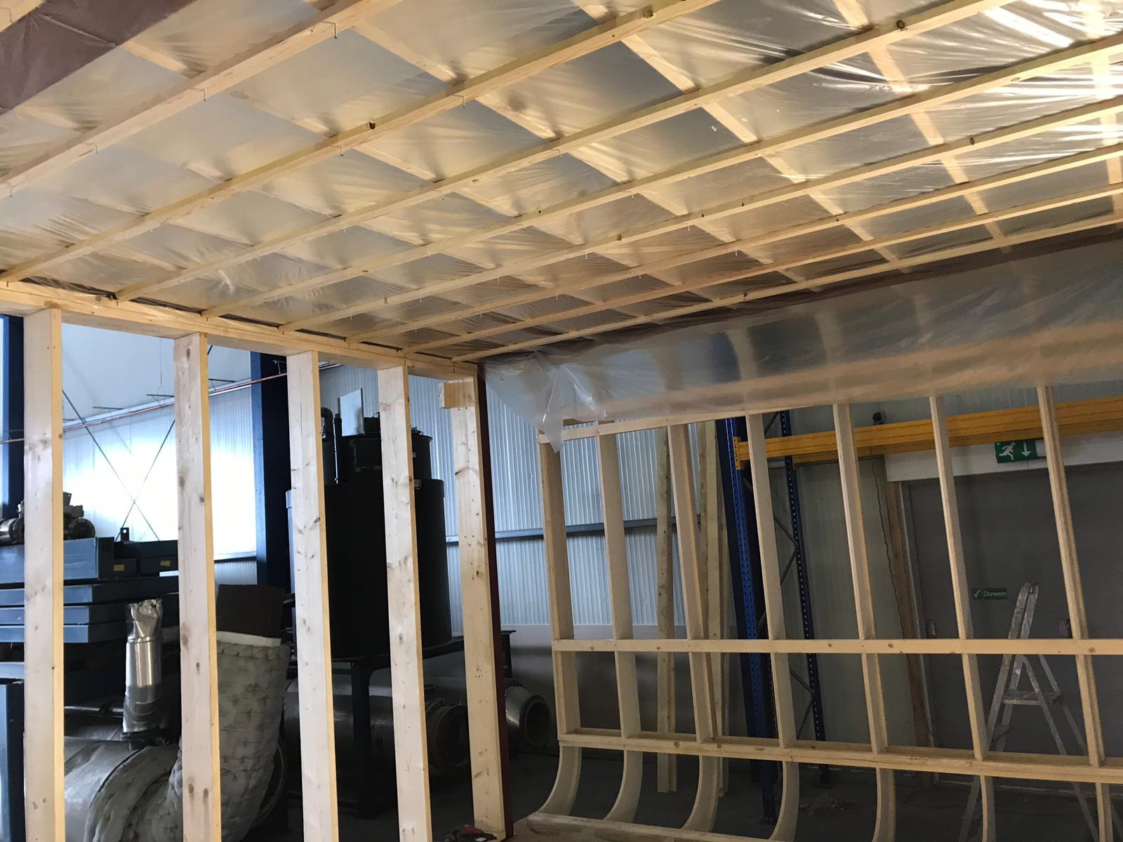 Drewniana konstrukcja ścian i sufitu w trakcie budowy, zabezpieczona folią, widoczne elementy instalacji i regały magazynowe w tle.