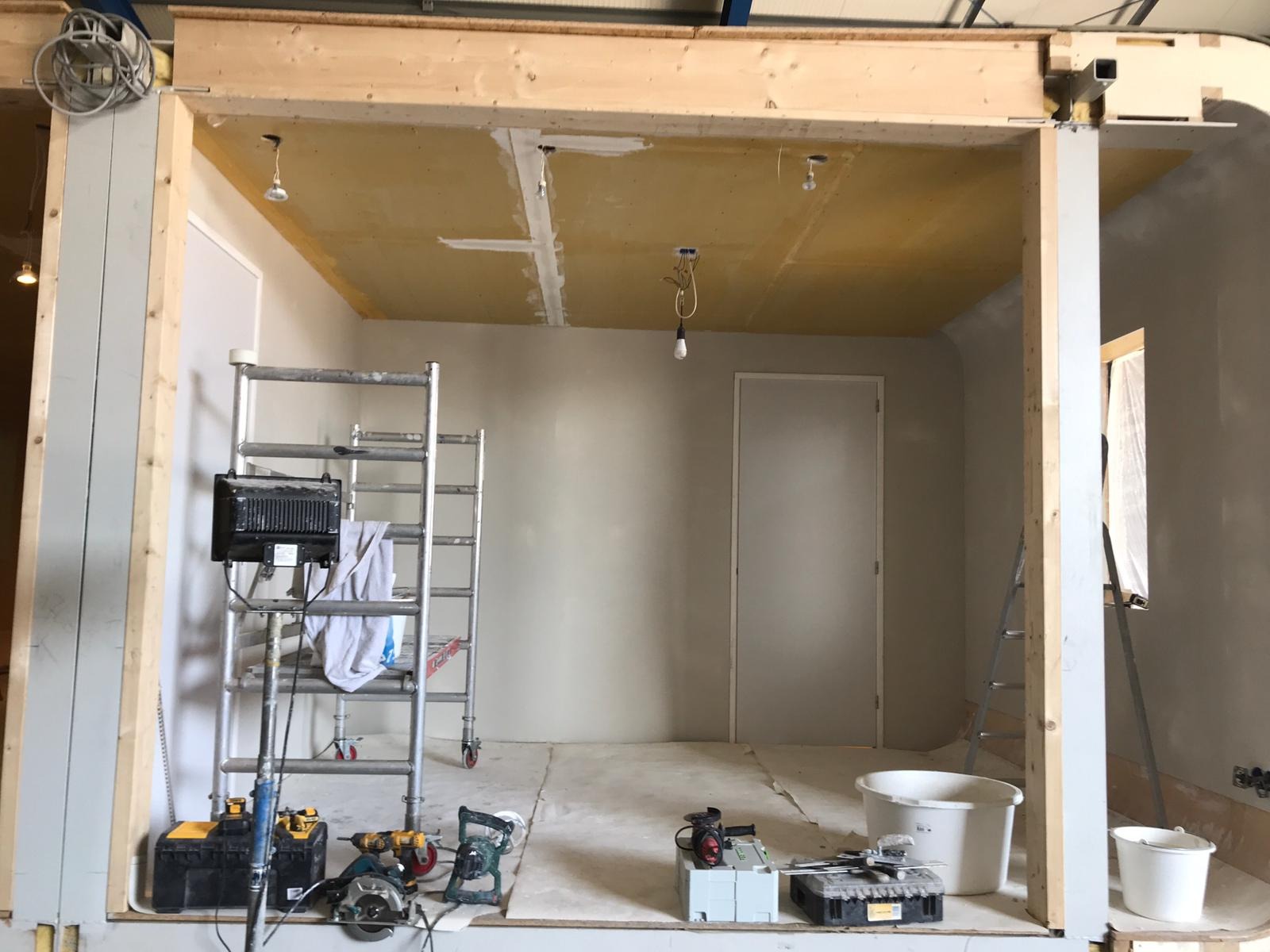 Wnętrze w trakcie remontu: ściany przygotowane do malowania, sufit z widocznymi łączeniami płyt gipsowych, drabina, narzędzia budowlane i lampa halogenowa.