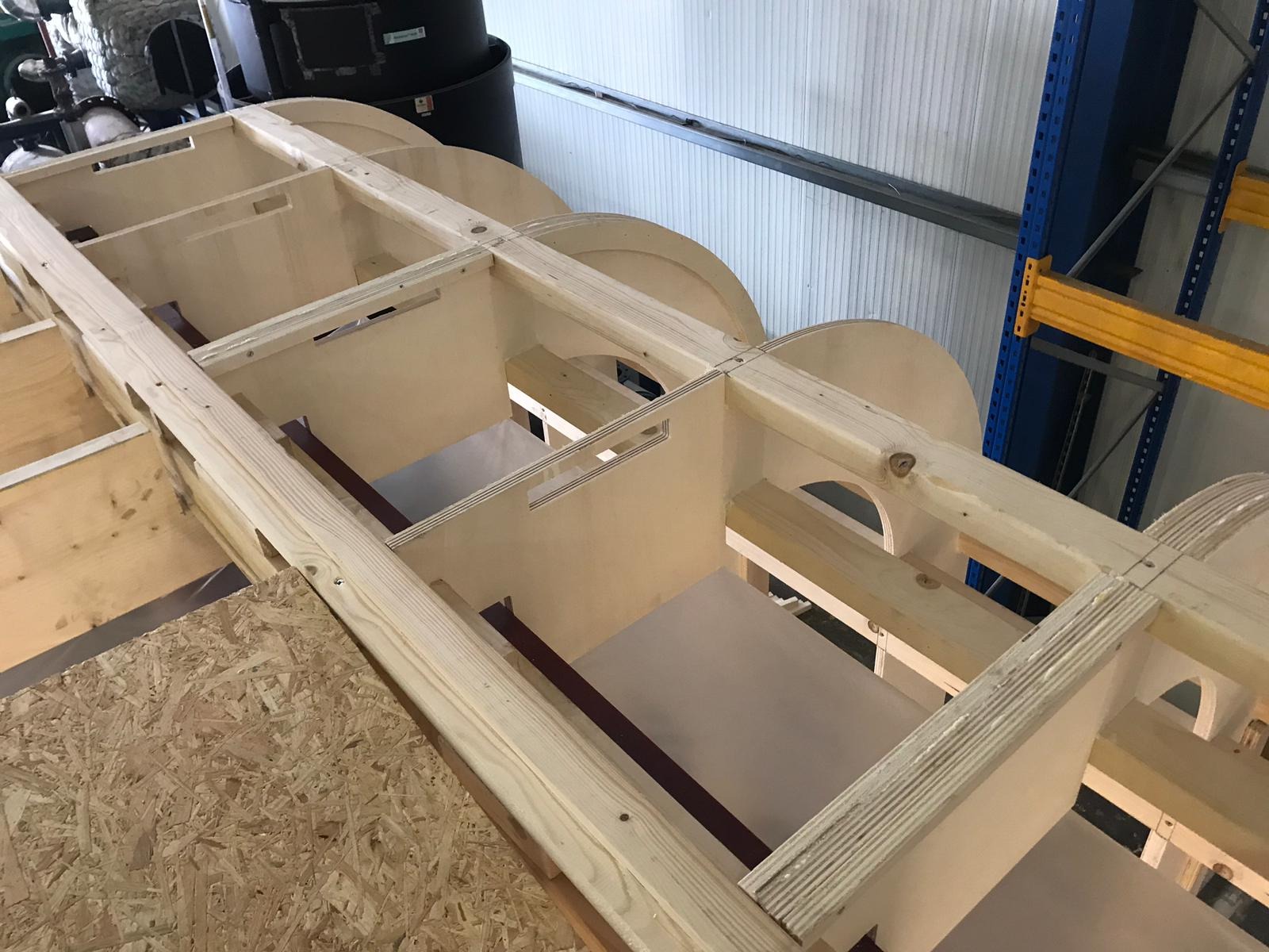 Drewniana konstrukcja szkieletowa z jasnego drewna, częściowo złożona, z widocznymi elementami łączącymi i okrągłymi wycięciami, umieszczona w hali produkcyjnej z regałami w tle.