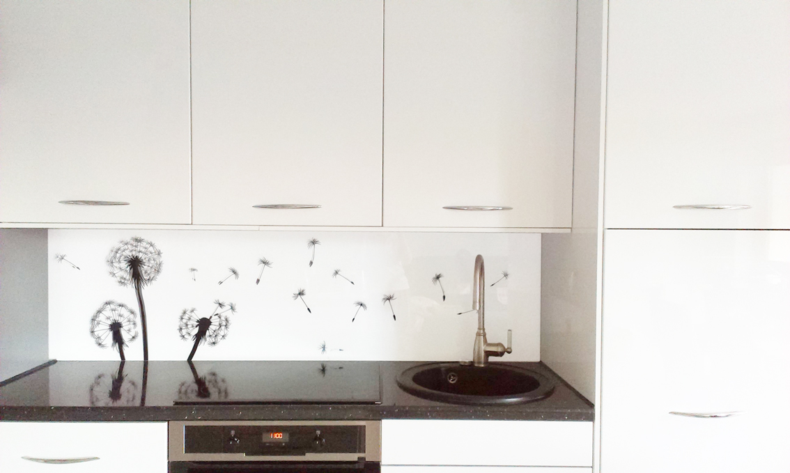 Biała kuchnia z panelami szklanymi z motywem dmuchawców nad blatem, zrealizowana w Andrychowie. Widoczne białe szafki, czarny blat i zlewozmywak.