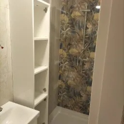 Nowoczesna łazienka z kabiną prysznicową, wzorzystymi płytkami i białą szafką wiszącą z otwartymi półkami, widoczna umywalka.