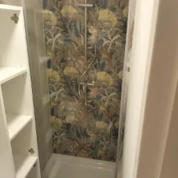 Kabina prysznicowa z deszczownicą i baterią, wykończona płytkami w motywy roślinne; obok otwarta szafka łazienkowa z białymi półkami.