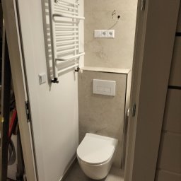 Bau Kris - Nowoczesna toaleta podwieszana z białym panelem spłukującym, grzejnik drabinkowy i gniazdka elektryczne na beżowych płytkach w łazience w Lwówku Śląskim. Widoczne niedokończone wykończenie.