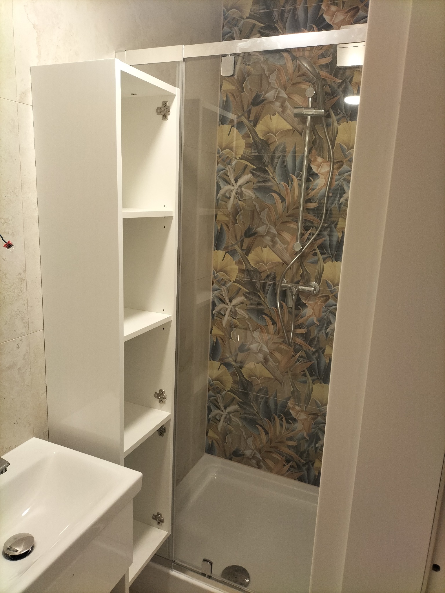 Nowoczesna łazienka z kabiną prysznicową, wzorzystymi płytkami i białą szafką wiszącą z otwartymi półkami, widoczna umywalka.