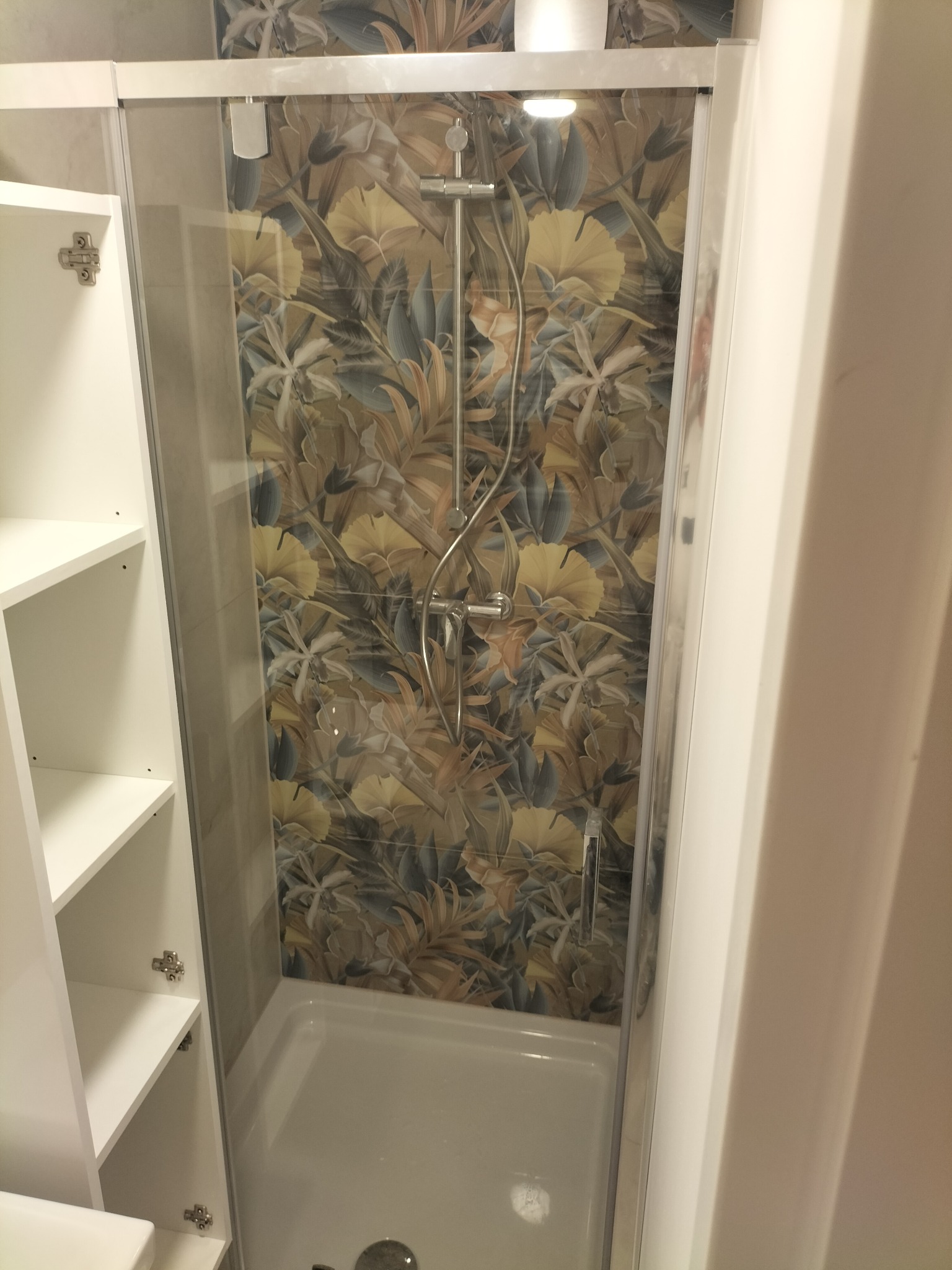 Kabina prysznicowa z deszczownicą i baterią, wykończona płytkami w motywy roślinne; obok otwarta szafka łazienkowa z białymi półkami.