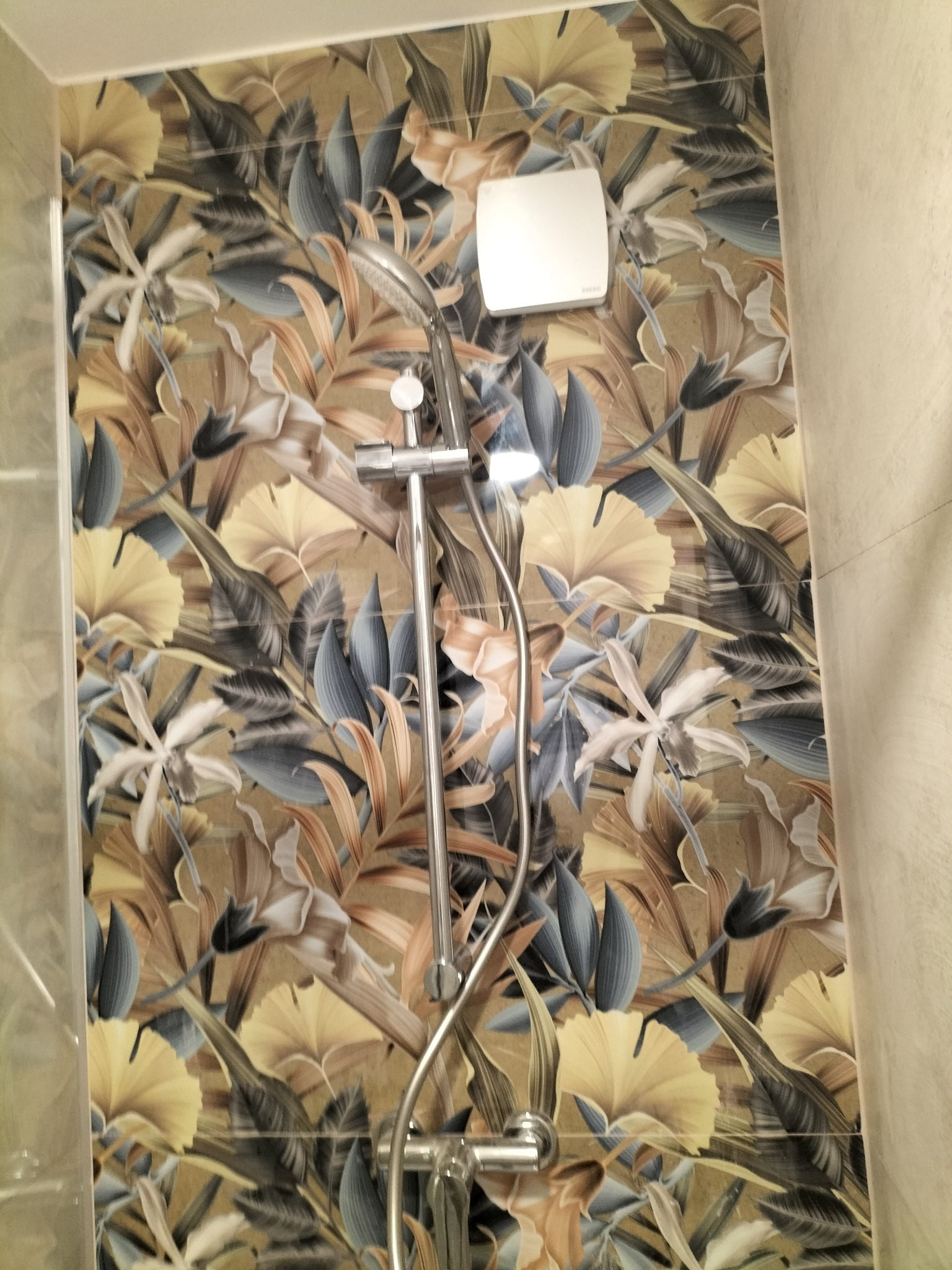 Nowoczesna kabina prysznicowa z baterią i deszczownicą na tle ściany wyłożonej płytkami z motywem tropikalnych liści w odcieniach beżu, żółci i szarości, widoczny także biały wentylator.