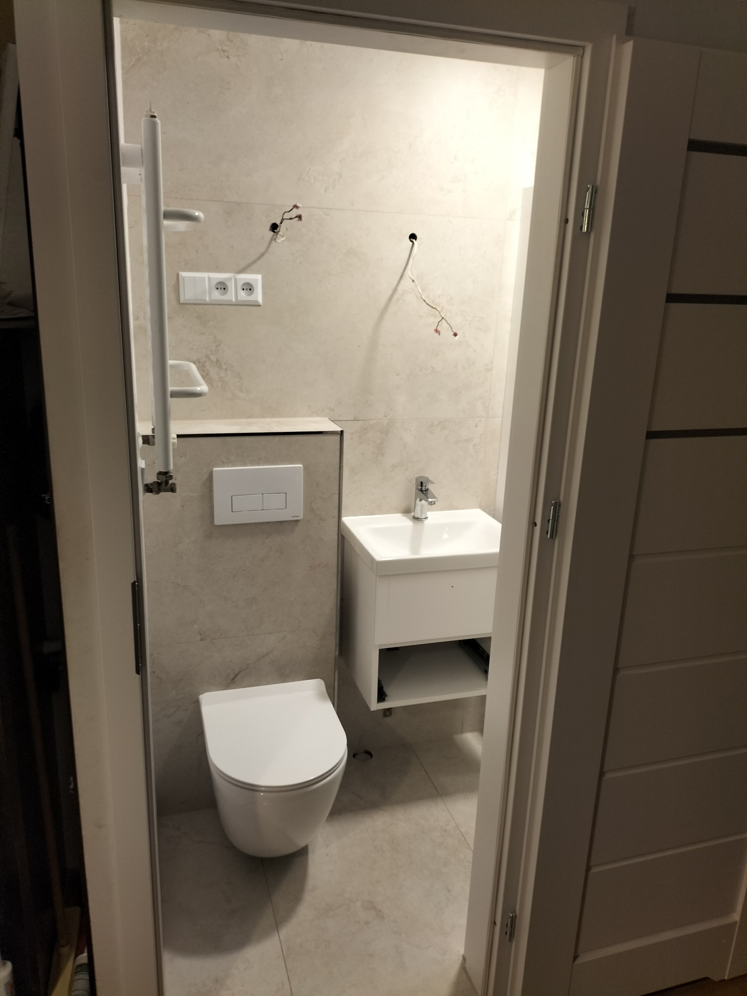 Widok przez otwarte drzwi na łazienkę w trakcie remontu: podwieszana toaleta, umywalka z szafką, grzejnik drabinkowy, płytki beżowe, wystające kable ze ściany.