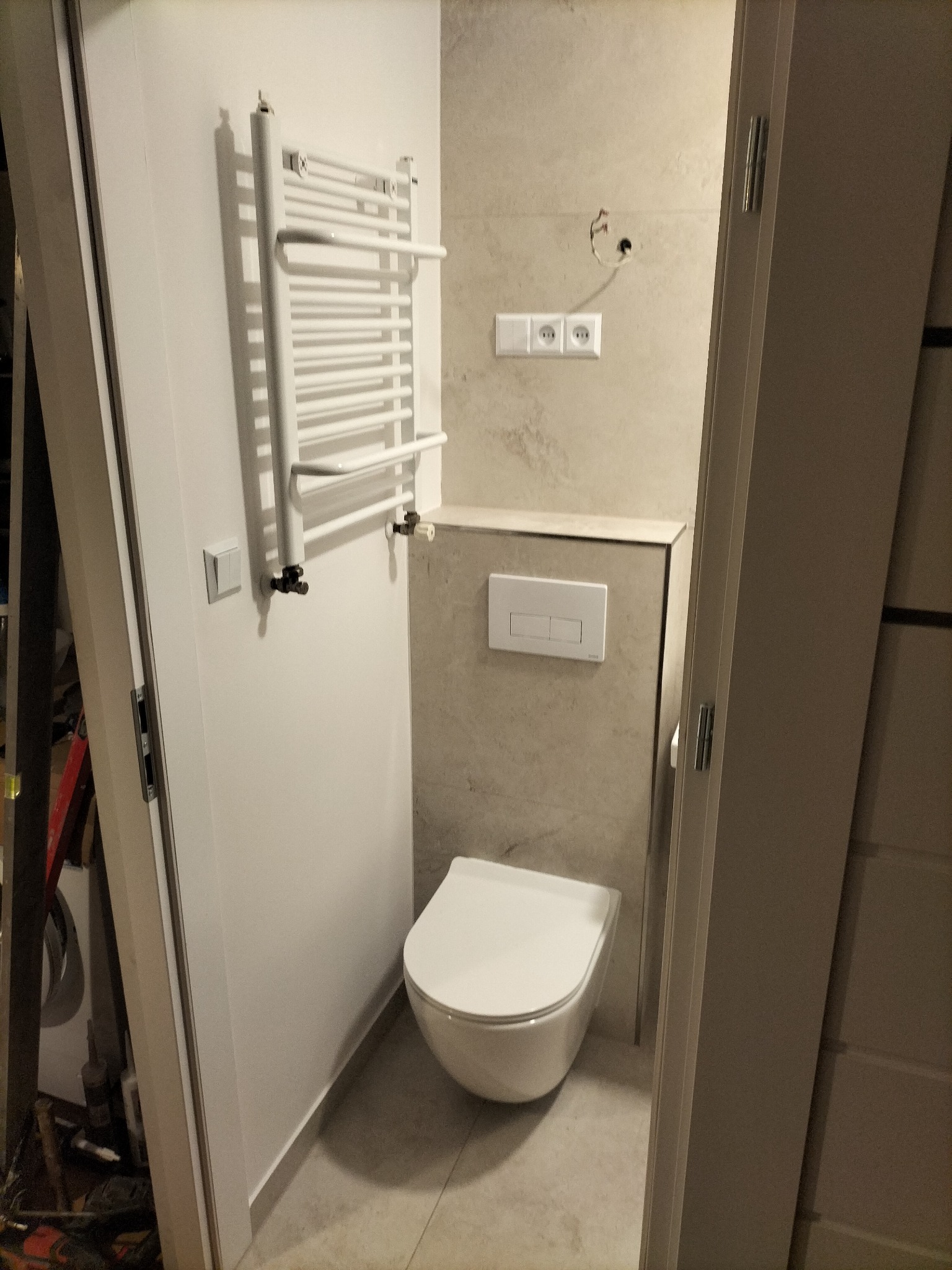 Nowoczesna toaleta podwieszana z białym panelem spłukującym, grzejnik drabinkowy i gniazdka elektryczne na beżowych płytkach w łazience w Lwówku Śląskim. Widoczne niedokończone wykończenie.