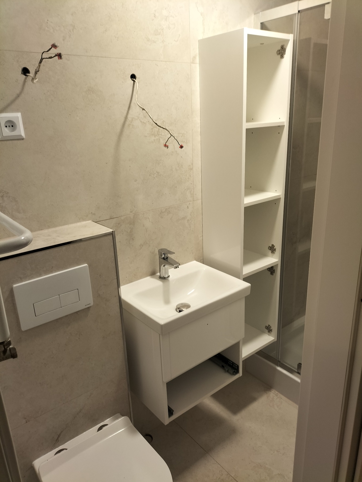 Nowoczesna łazienka w trakcie wykańczania: biała umywalka z szafką, toaleta podwieszana, wysoka szafka i kabina prysznicowa, widoczne niedokończone instalacje elektryczne na ścianie z beżowych płytek.