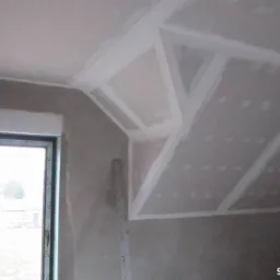 Skośny sufit poddasza z zamontowaną płytą kartonowo-gipsową, widoczne łączenia płyt zabezpieczone taśmą i szpachlą, okno w tle.