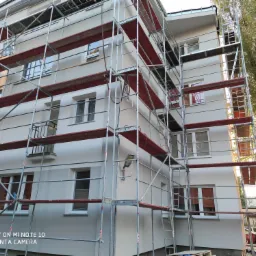 Wielopiętrowy budynek mieszkalny w trakcie prac elewacyjnych, otoczony rusztowaniami z czerwonymi podestami i świeżo pomalowaną, jasną elewacją, widoczne okna z roletami.