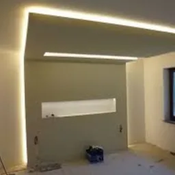 Wnętrze w trakcie prac wykończeniowych z podwieszonym sufitem i oświetleniem LED, widoczne narzędzia i surowa podłoga.