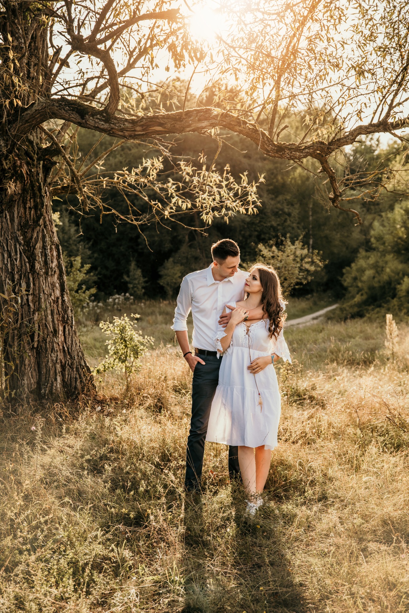 Zakochana para w objęciach pod rozłożystym drzewem w złotym świetle zachodzącego słońca na łące w okolicach Poznania. Kobieta w białej sukience trzyma w ręku ozdobę z piór.