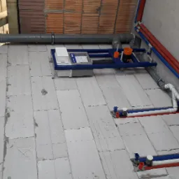 Sufit i podłoga w trakcie budowy z widocznymi szarymi rurami kanalizacyjnymi, niebieskimi i czerwonymi rurami instalacji wodnej oraz stelażem podtynkowym WC z widocznym zbiornikiem i elementami...