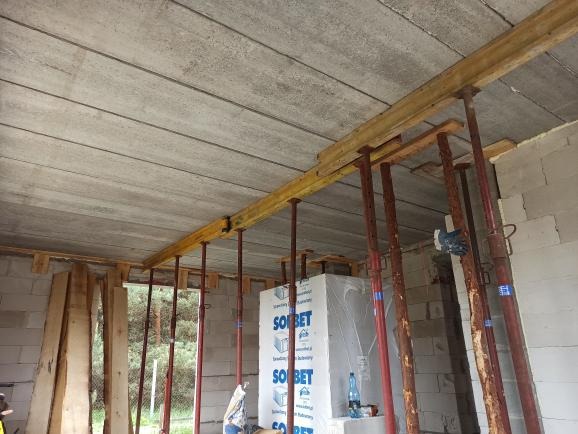 Wnętrze budowy domu w trakcie wznoszenia ścian z betonu komórkowego, z widocznym stropem prefabrykowanym podpartym stemplami stalowymi i drewnianymi belkami, bloczki Solbet w tle.
