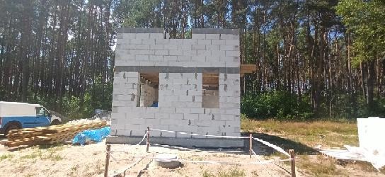 Wznoszone ściany domu z betonu komórkowego, widoczne otwory okienne, na pierwszym planie liny wyznaczające teren budowy, w tle las i samochód dostawczy.