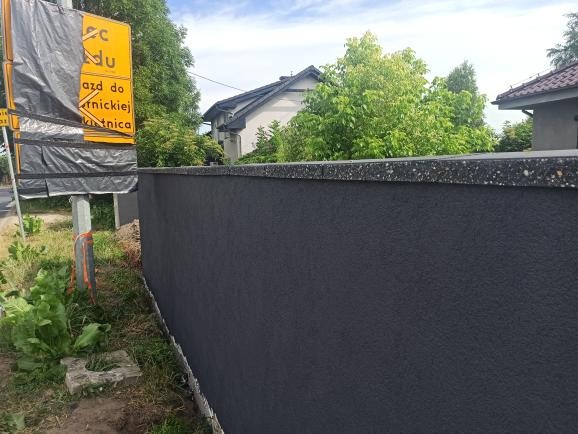 Nowo otynkowane, grafitowe ogrodzenie posesji z szarą, nakrapianą czapą, widoczne z boku, z fragmentem zasłoniętego znaku drogowego w tle.