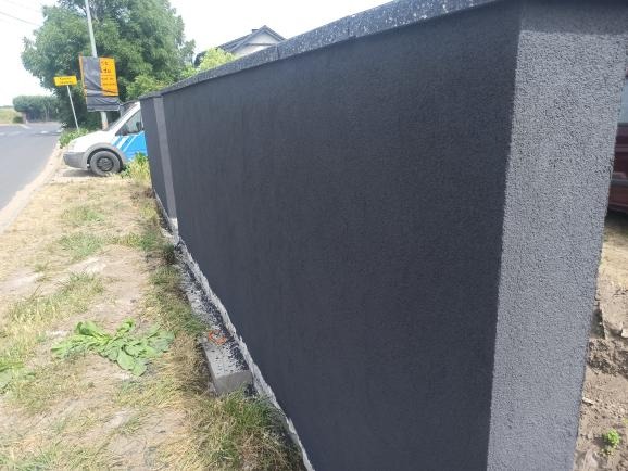 Ocieplony, pomalowany na ciemno szary mur ogrodzeniowy z widoczną fakturą, z fragmentem zaparkowanego samochodu w tle, oraz trawa i chwasty przy podstawie muru.