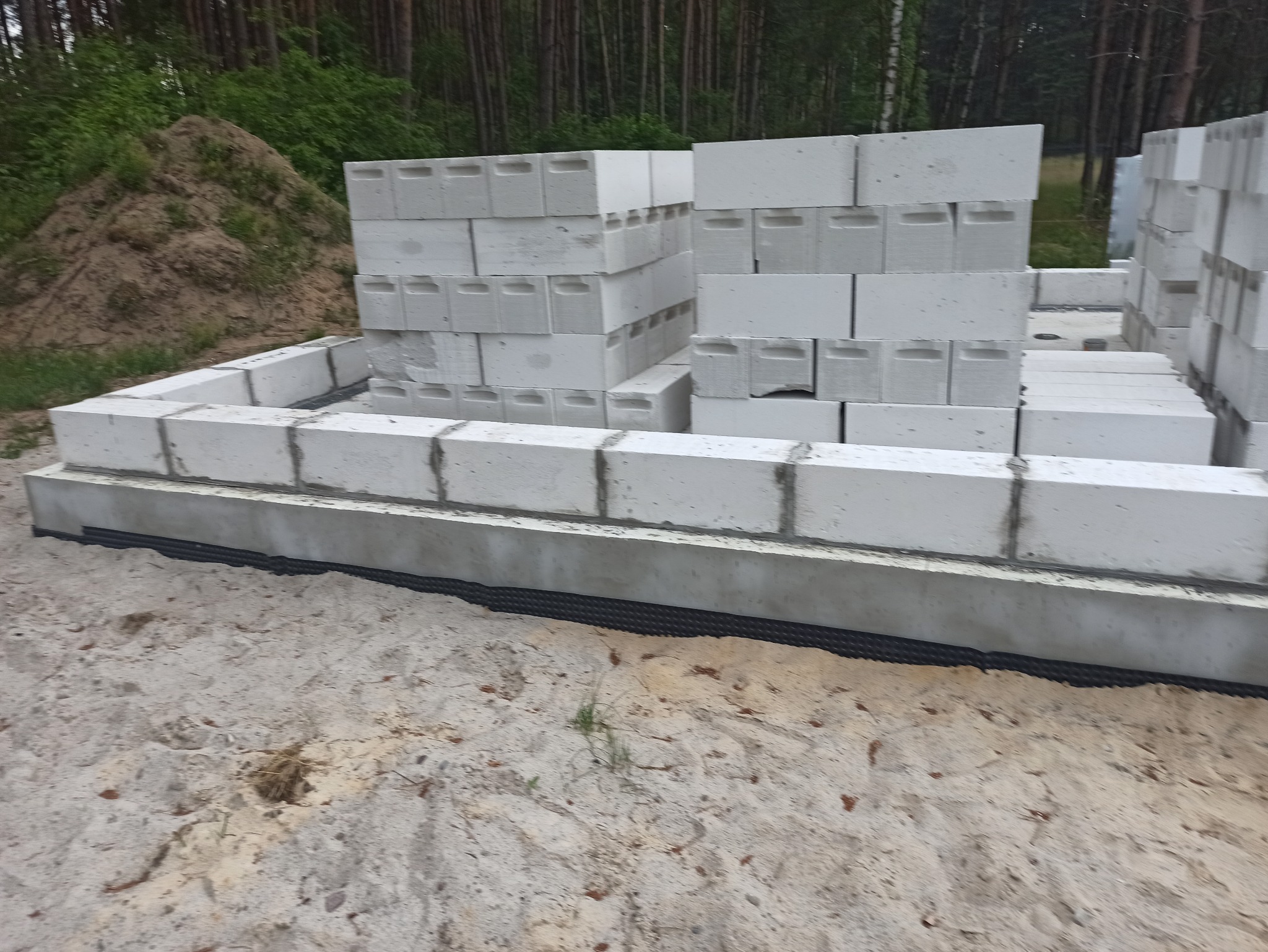 Wznoszone fundamenty domu z bloczków betonowych na piasku, widoczna czarna izolacja pozioma i stos bloczków w tle, teren budowy.
