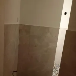 Wykończona łazienka z beżowymi płytkami imitującymi marmur na ścianach i wzorzystymi płytkami na podłodze, widoczne fragmenty ścian i sufitu w kolorze białym, brak armatury.