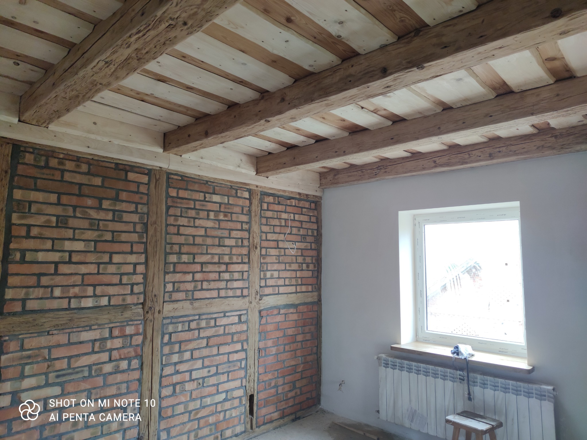 Wnętrze w trakcie prac wykończeniowych: ceglana ściana z drewnianą konstrukcją, drewniany sufit z belkami, okno z parapetem i grzejnikiem, mały drewniany stołek.
