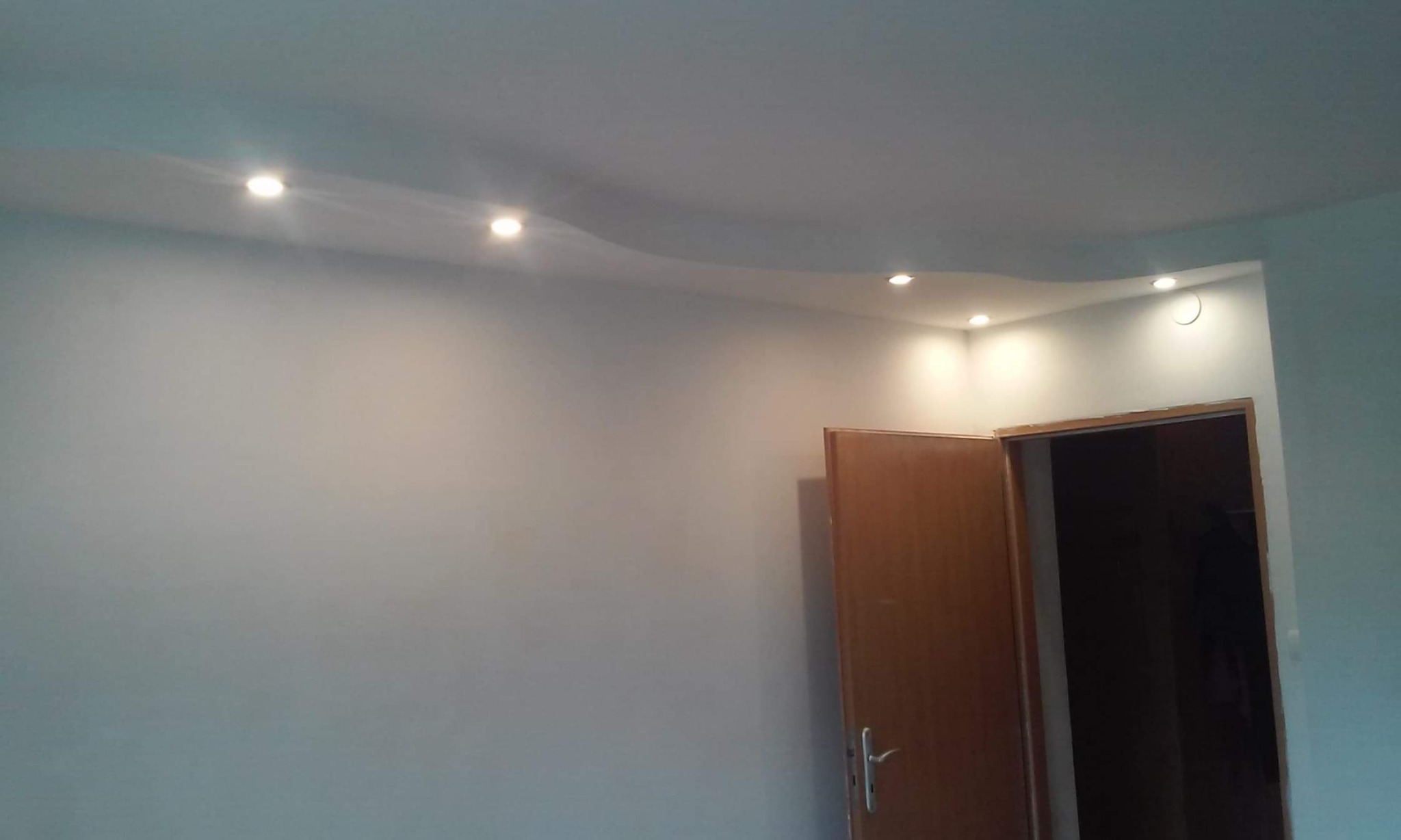 Świeżo pomalowane wnętrze z gładką, jasną ścianą i sufitem z wbudowanymi punktowymi światłami LED oraz częściowo otwartymi drzwiami z widocznym wnętrzem pomieszczenia za nimi.