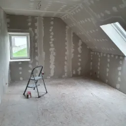 Poddasze w trakcie przygotowania do wykończenia, widoczne płyty karton-gips z nałożoną masą szpachlową, drabina aluminiowa i okno dachowe.