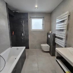 Nowoczesna łazienka w odcieniach szarości z wanną, kabiną prysznicową typu walk-in z czarnymi płytkami imitującymi marmur, podwieszaną toaletą i grzejnikiem drabinkowym.