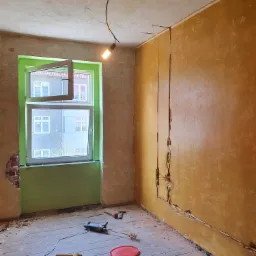 Pokój w trakcie odnawiania: surowe ściany z żółtą farbą, odkryte przewody elektryczne w bruzdach, otwarte okno z zieloną ramą, narzędzia malarskie na podłodze z desek, wisząca żarówka