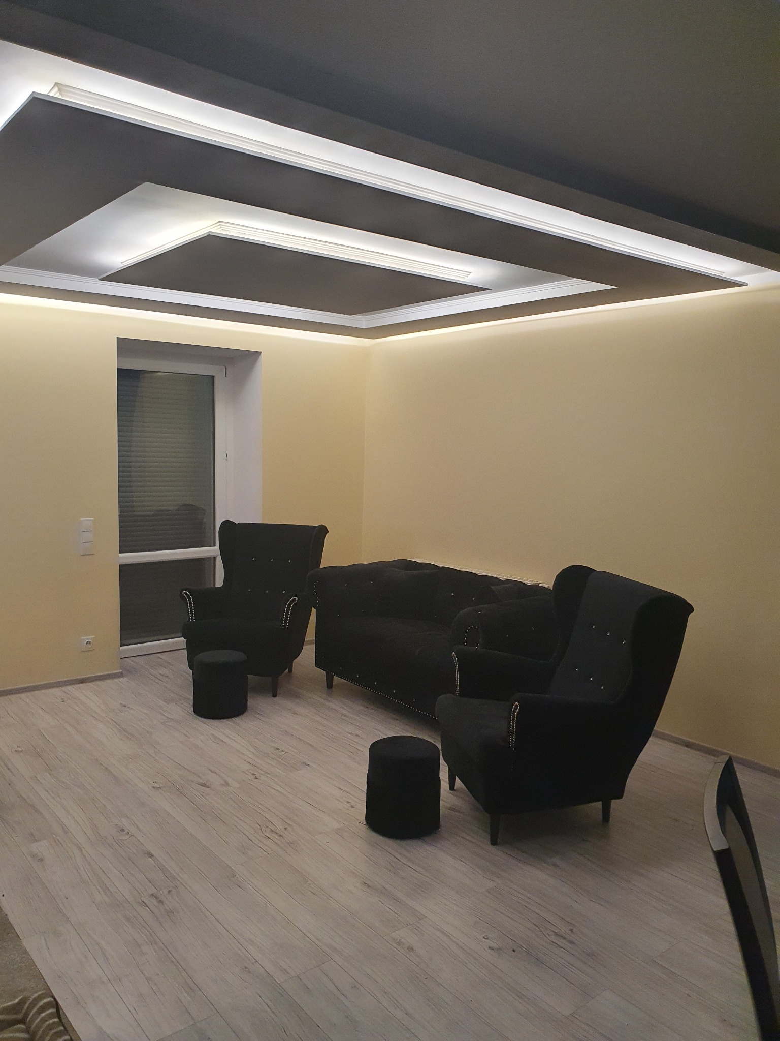 Salon z czarnymi meblami w stylu glamour: sofa, dwa fotele typu uszak i pufy, oświetlenie sufitowe LED w geometrycznej zabudowie, jasne ściany i podłoga imitująca drewno.