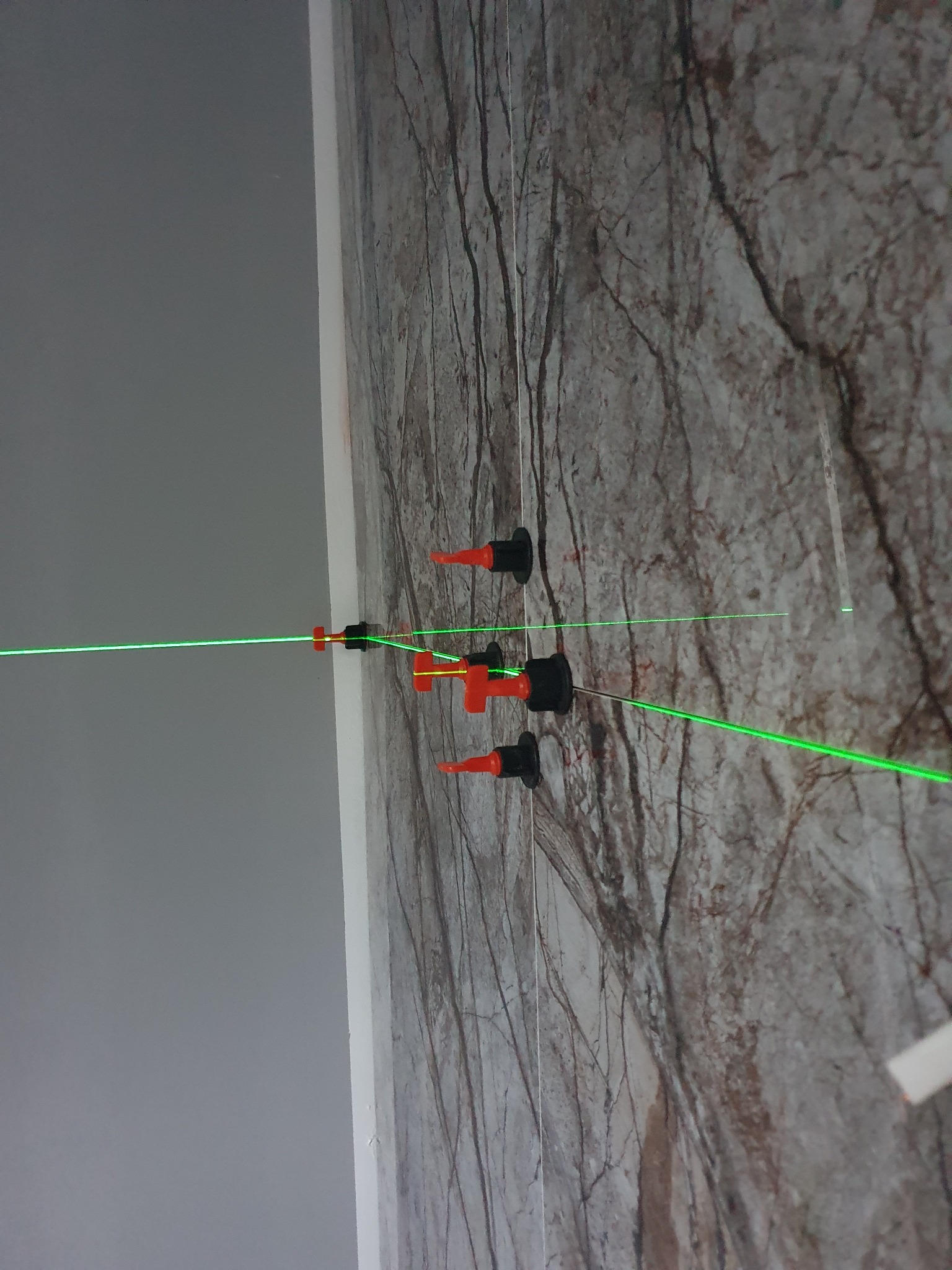 Układanie płytek imitujących marmur na ścianie, widoczny laserowy poziom i system poziomowania płytek z czerwonymi klinami i czarnymi podstawkami.