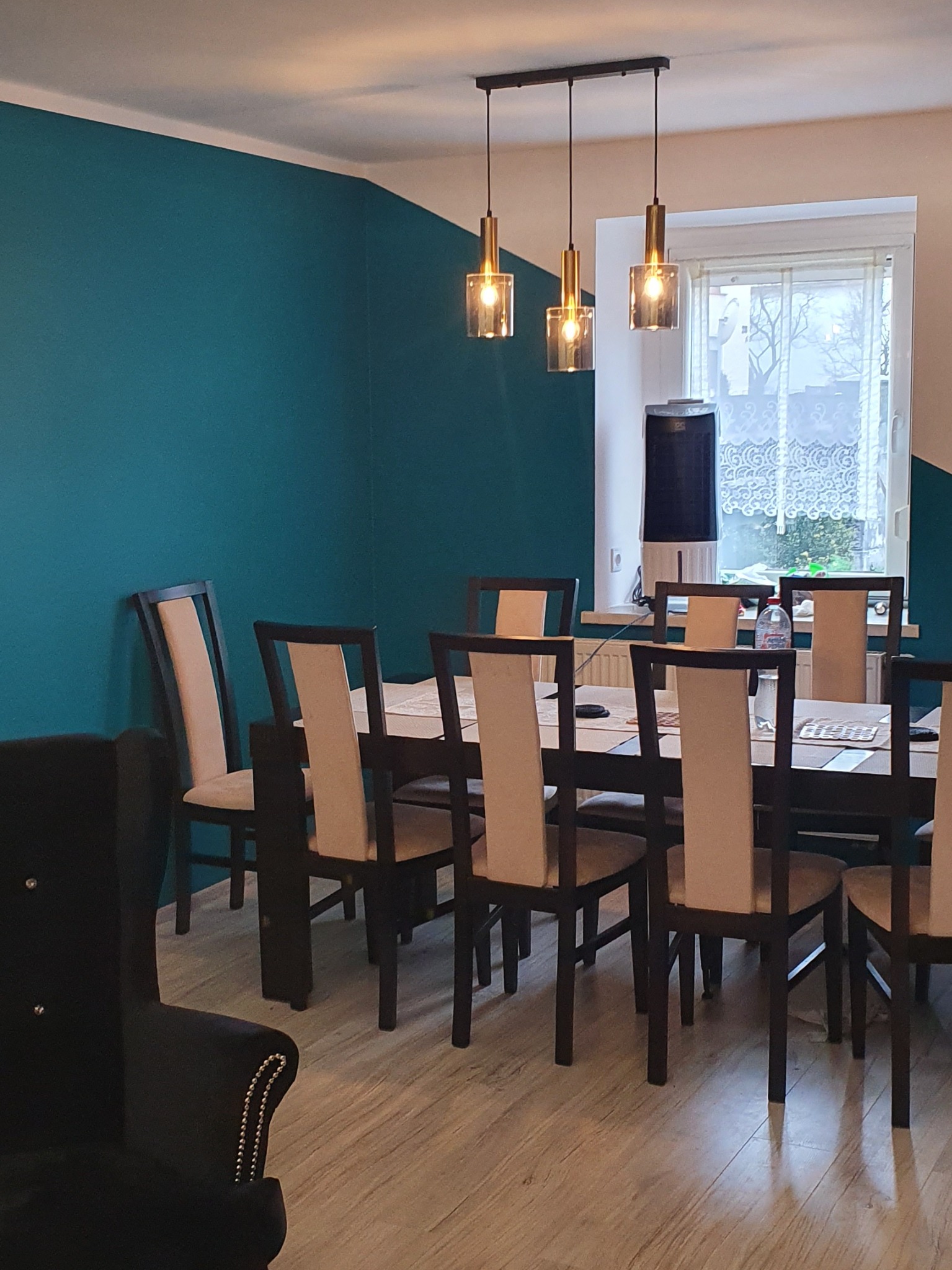 Pomieszczenie jadalne z turkusową ścianą, drewnianym stołem i krzesłami z beżowymi oparciami, oświetlone trzema wiszącymi lampami z kloszami w odcieniach złota i szkła.