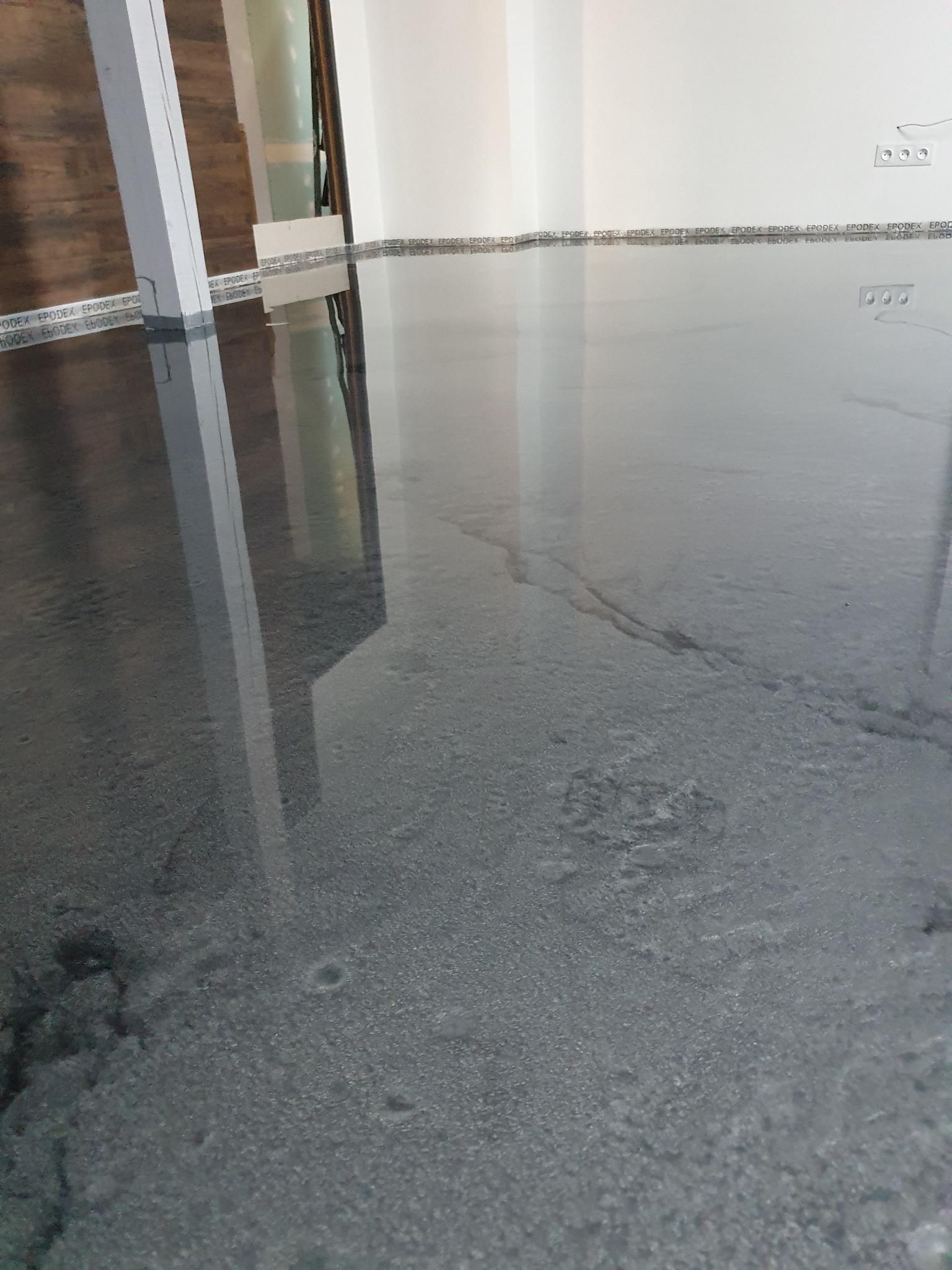 Błyszcząca, szara podłoga pokryta żywicą epoksydową, z widocznymi refleksami światła i taśmą ochronną przy ścianach, zabezpieczającą przed zabrudzeniem podczas aplikacji.