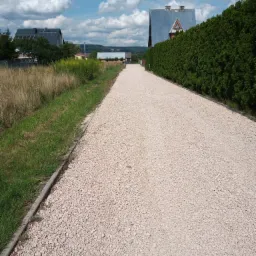Długa, prosta droga wysypana drobnym, jasnym żwirem, otoczona zielenią i tujami, prowadząca w kierunku zabudowań w oddali pod błękitnym niebem z chmurami.
