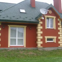 Dwukondygnacyjny dom z czerwoną elewacją, beżowymi detalami wokół okien i na narożach, zielonym dachem z dachówką i anteną satelitarną.