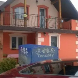 Dwupiętrowy dom z balkonem, pomalowany na pomarańczowo z białymi akcentami, częściowo zasłonięty przez przyczepkę z napisem 'TerazMy', widoczna kobieta w tle.