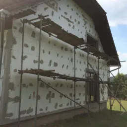 Dom w trakcie ocieplania styropianem, widoczne rusztowania i przyklejony styropian z pozostawionymi miejscami na klej, w tle zielone drzewa.