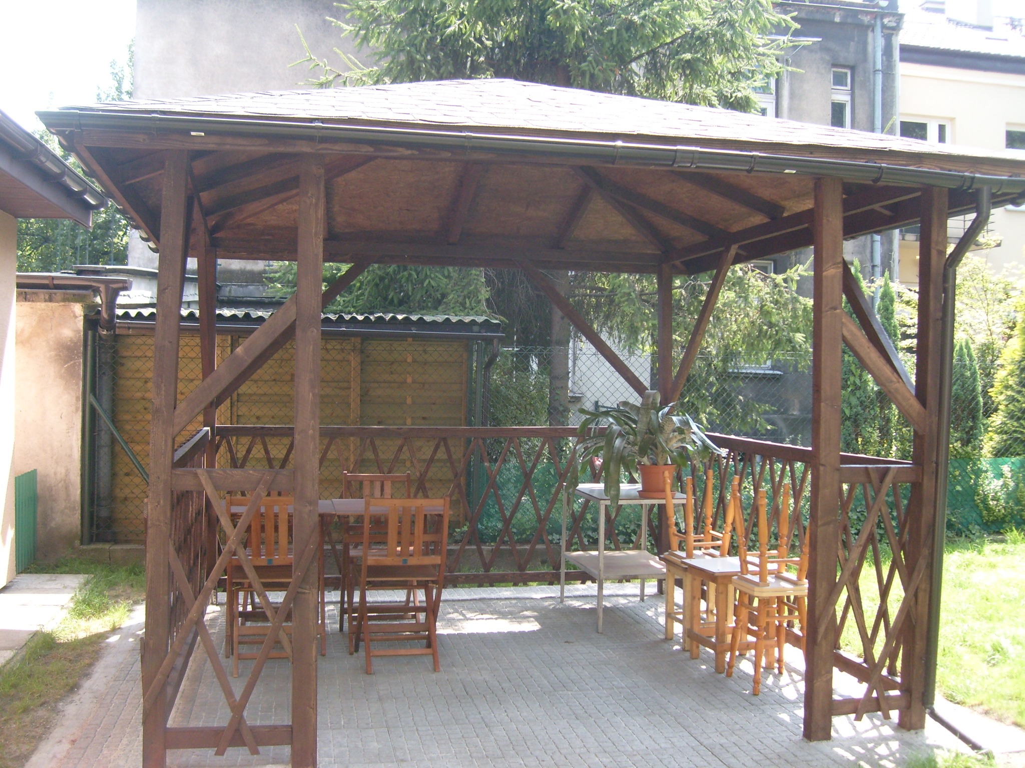 Drewniana altana ogrodowa z meblami, zadaszona bitumiczną dachówką, na wybrukowanym patio, z widoczną rośliną doniczkową na stoliku.