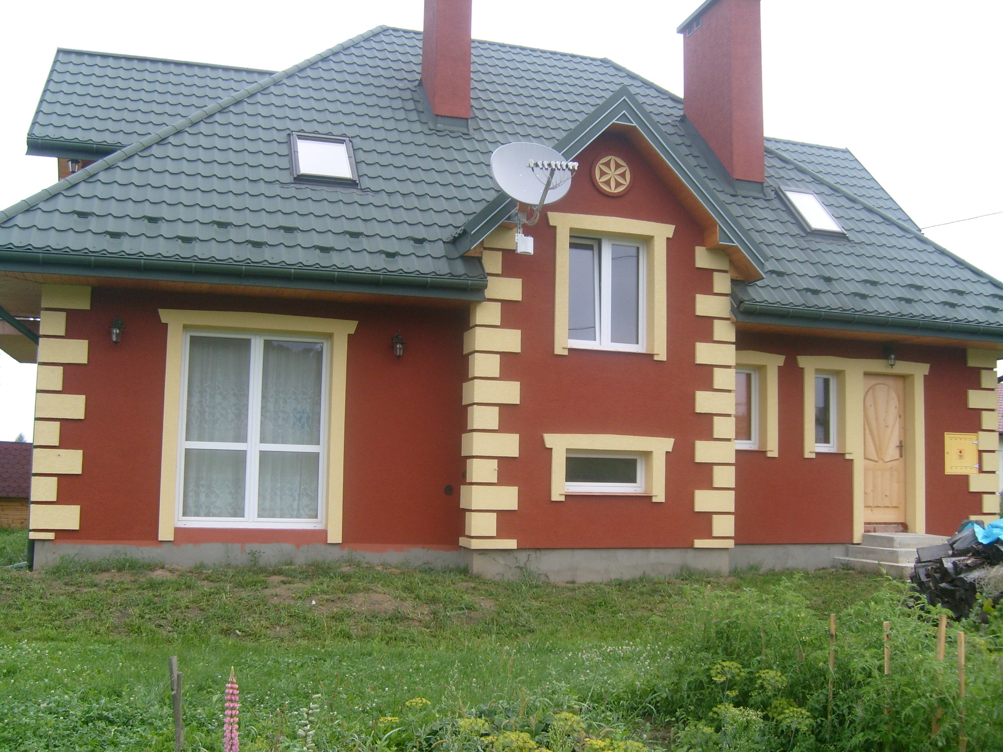 Dom z czerwoną elewacją ozdobioną żółtymi, wystającymi elementami, zielonym dachem z lukarnami i anteną satelitarną na szczycie.