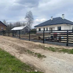 Czarna, pozioma konstrukcja ogrodzeniowa z metalowych profili na betonowej podmurówce, otaczająca posesję z dwoma domami o różnej architekturze, widoczna z perspektywy drogi gruntowej.