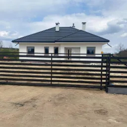 Nowoczesny dom jednorodzinny z czarną, poziomą bramą wjazdową, widoczne elementy konstrukcyjne i niezabezpieczone ściany zewnętrzne.