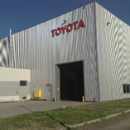 Duży budynek przemysłowy z blachy trapezowej w kolorze srebrnym z czerwonym logo Toyota, widoczny otwarty wjazd i drzwi wejściowe, na zewnątrz kostka brukowa i trawnik.